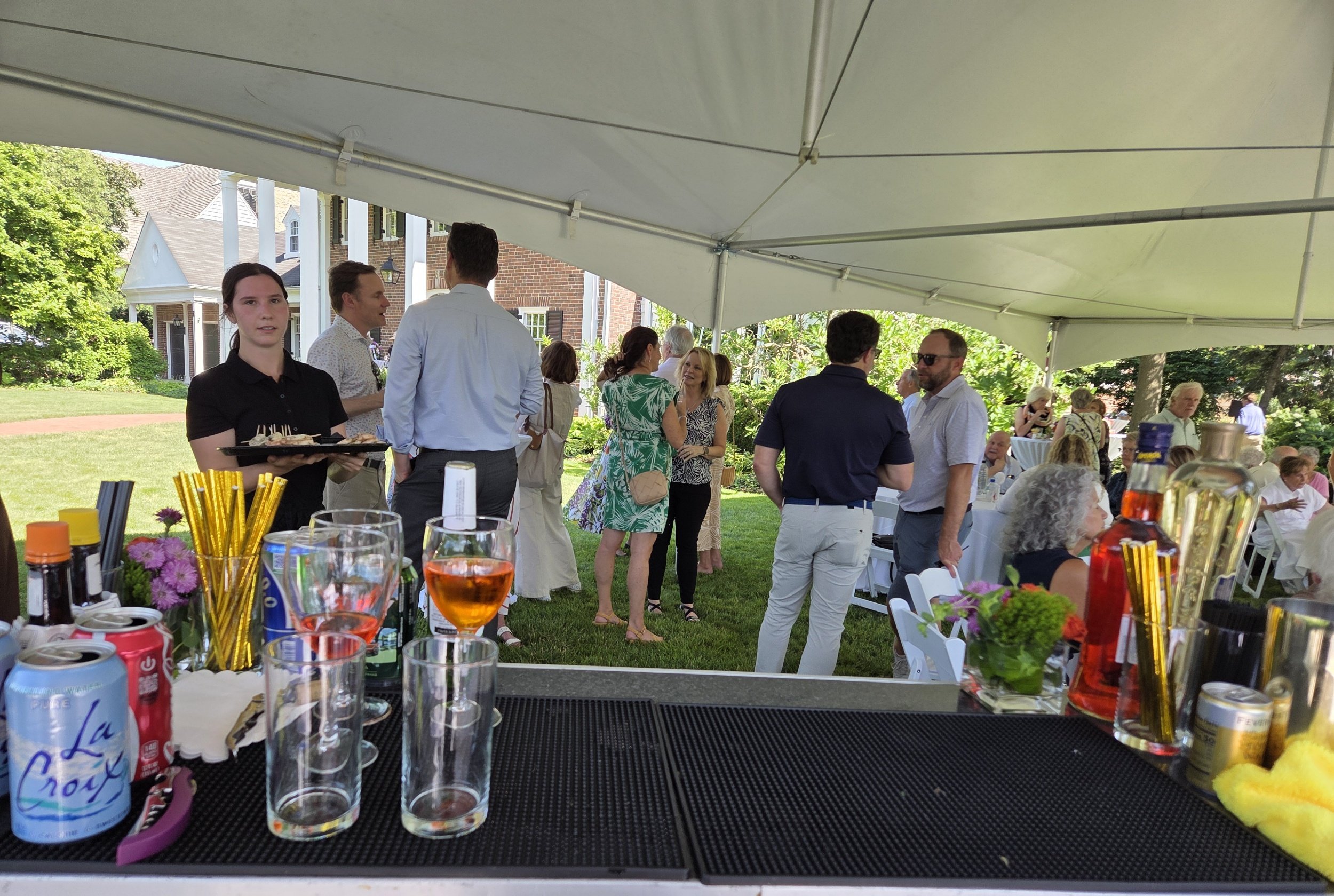 People socializing at an outdoor gathering under a white canopy with a table of drinks and flowers in the foreground.