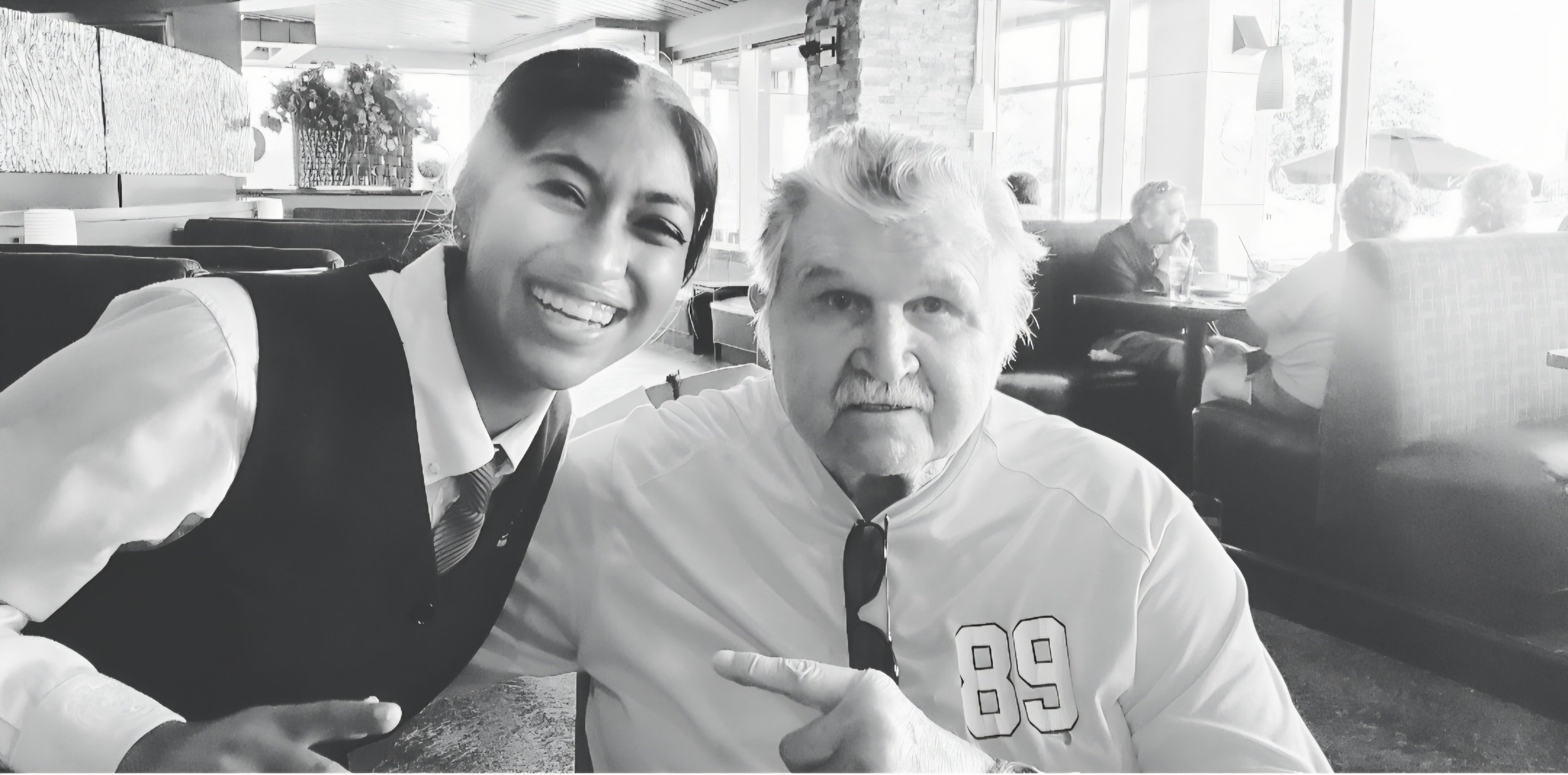 A young woman with dark hair, smiling widely, posing with an older man with white hair and a mustache, pointing at him while sitting in a restaurant or cafe. They are indoors with other patrons and large windows in the background.