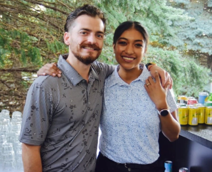 A man and woman smiling and standing close together outdoors, with trees in the background and drinks on a table behind them.