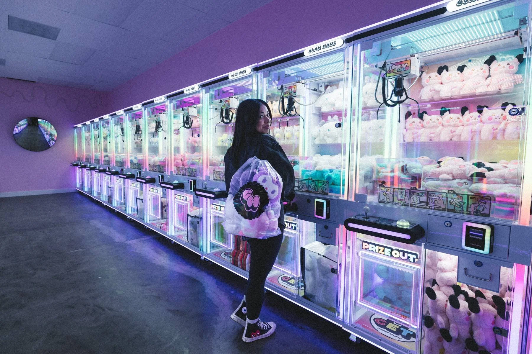 A woman with dark hair, wearing a black jacket and patterned sneakers, stands in front of a row of colorful claw machines filled with plush stuffed animals, in an arcade with purple walls and neon lighting.