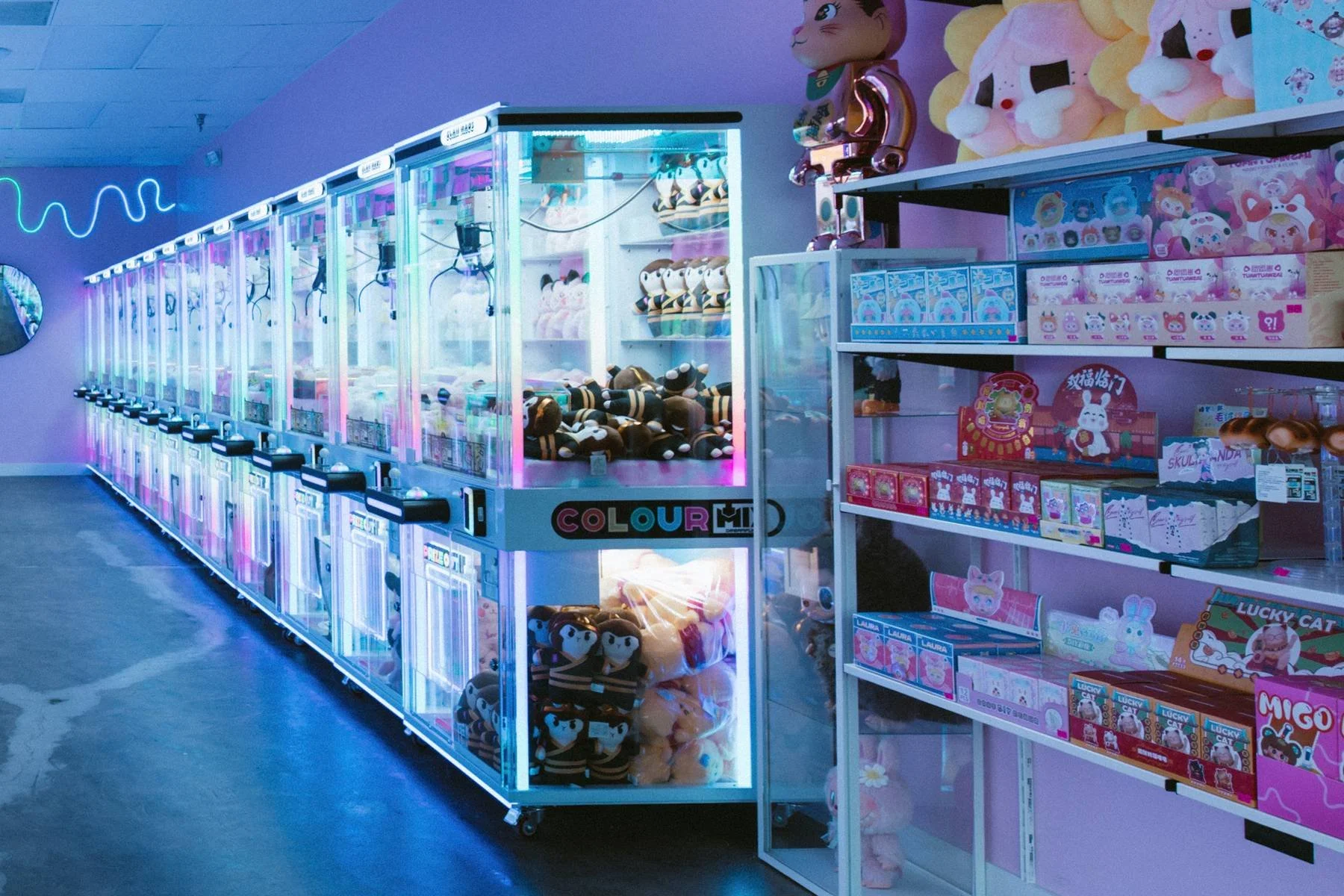 colorful claw machine filled with plush toys, next to a shelf with toy boxes and plushies in a store.