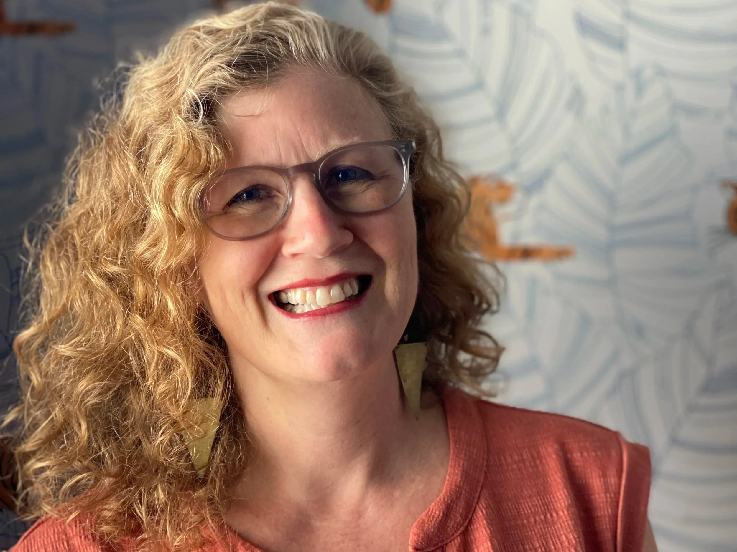 Close-up of a smiling woman with curly blonde hair, wearing glasses and a reddish-brown top, against a background of a wall with patterned tiles.