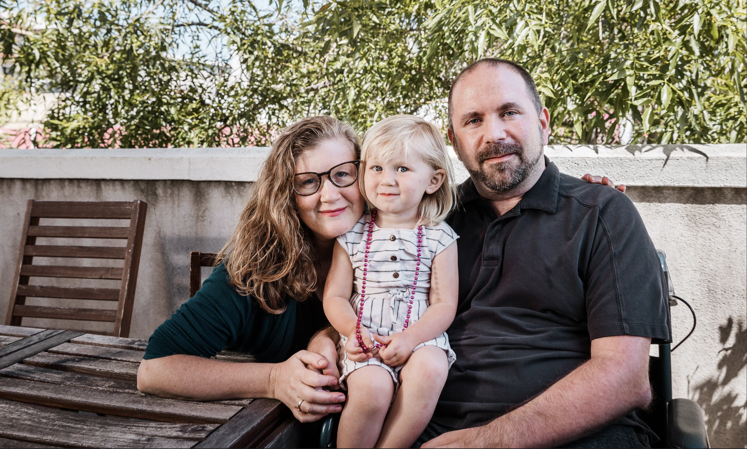 A family of three sitting outside on a wooden table with green foliage in the background. The woman with long, curly red hair and glasses is next to a young girl with blonde hair, and an adult man with a beard is sitting beside her.