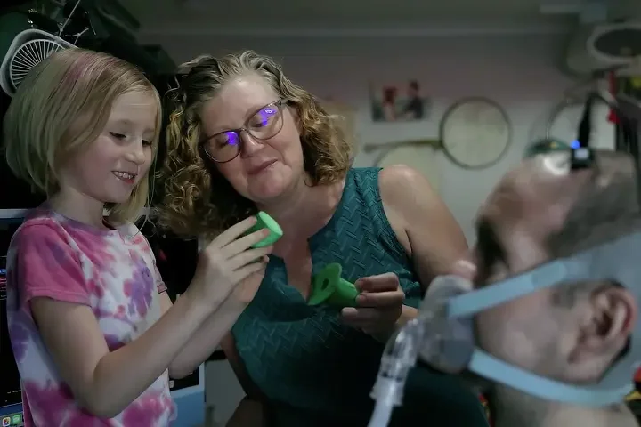 A young girl, a woman, and a baby in a hospital room. The girl and woman are smiling and looking at a device in the girl's hand. The baby is on a hospital bed with medical equipment.