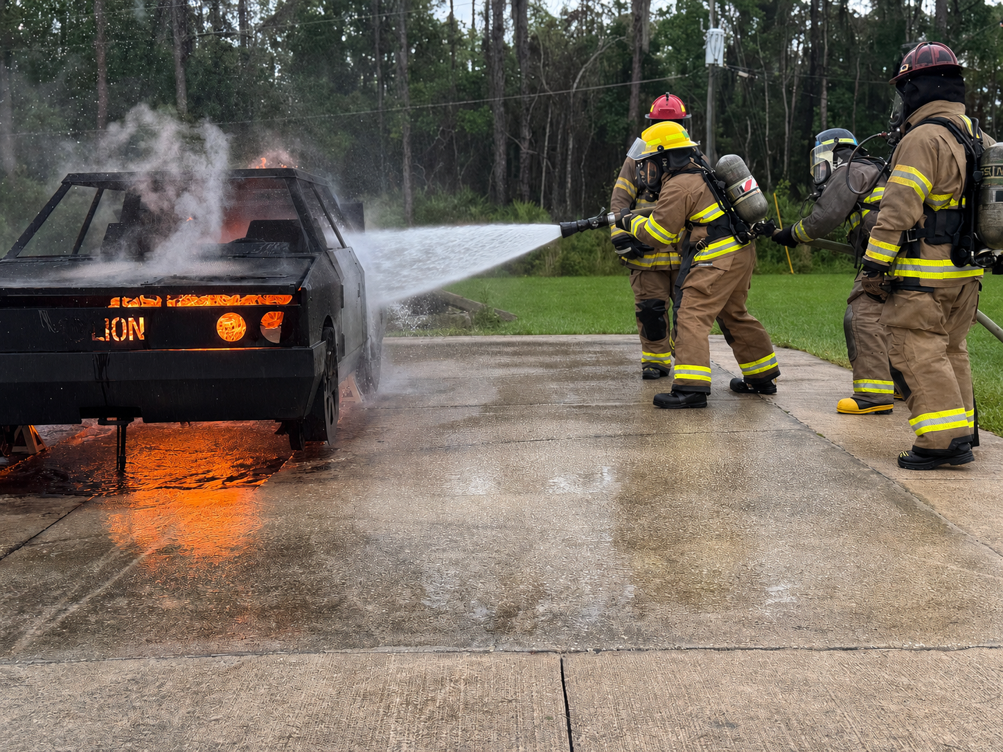 Firefighters extinguishing a car fire on a driveway, with the car shaped like a toy or model car, surrounded by trees.