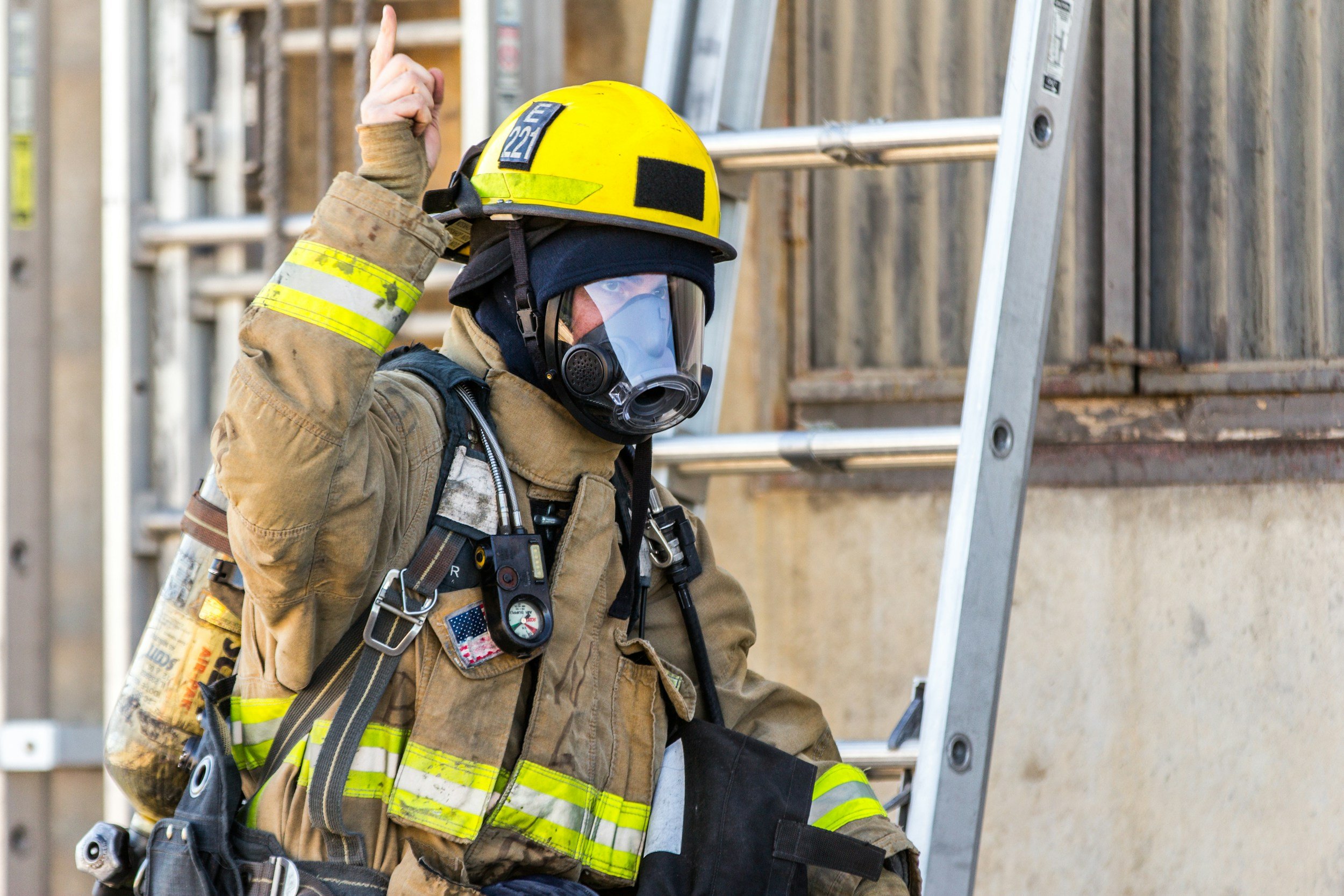 Firefighter wearing a yellow helmet, protective gear, and a gas mask, raising one finger while standing near a ladder outside a building.