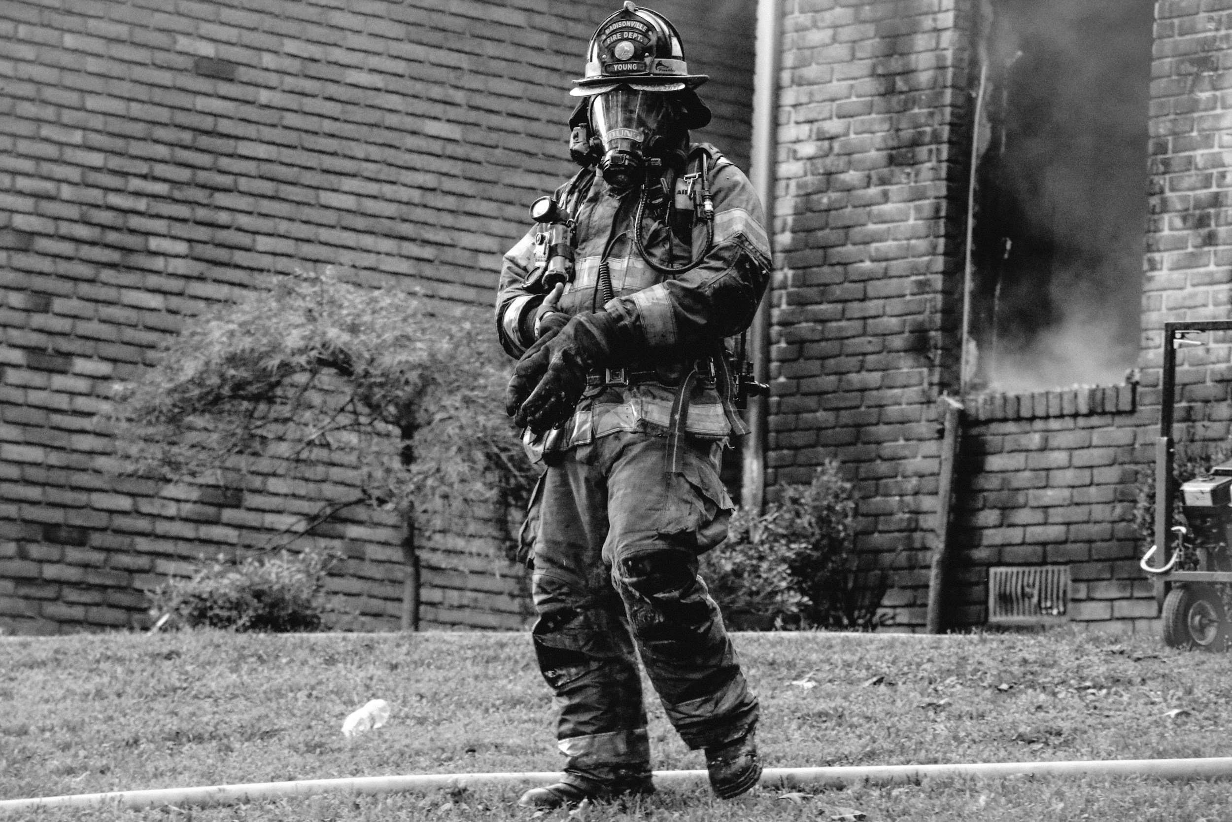 A firefighter in full gear, including a helmet, mask, and gloves, standing outside a building with smoke coming from a window.