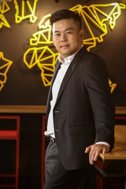 A young man in a dark suit and white shirt, standing in a modern, warmly lit room with a black wall decorated with yellow geometric world map outlines.