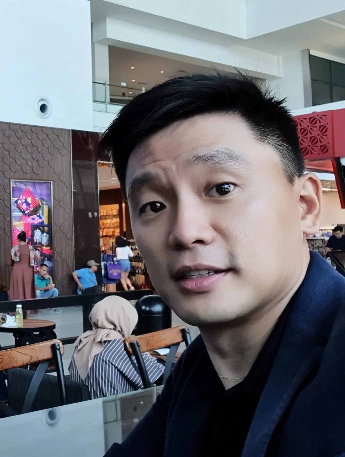 A man taking a selfie in a busy, modern shopping mall or food court, with other patrons and store displays in the background.