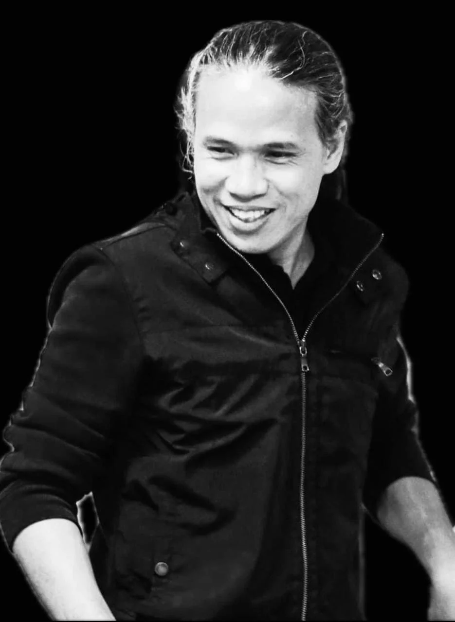 Black and white photo of a smiling man with long hair, wearing a dark jacket with a zipper.