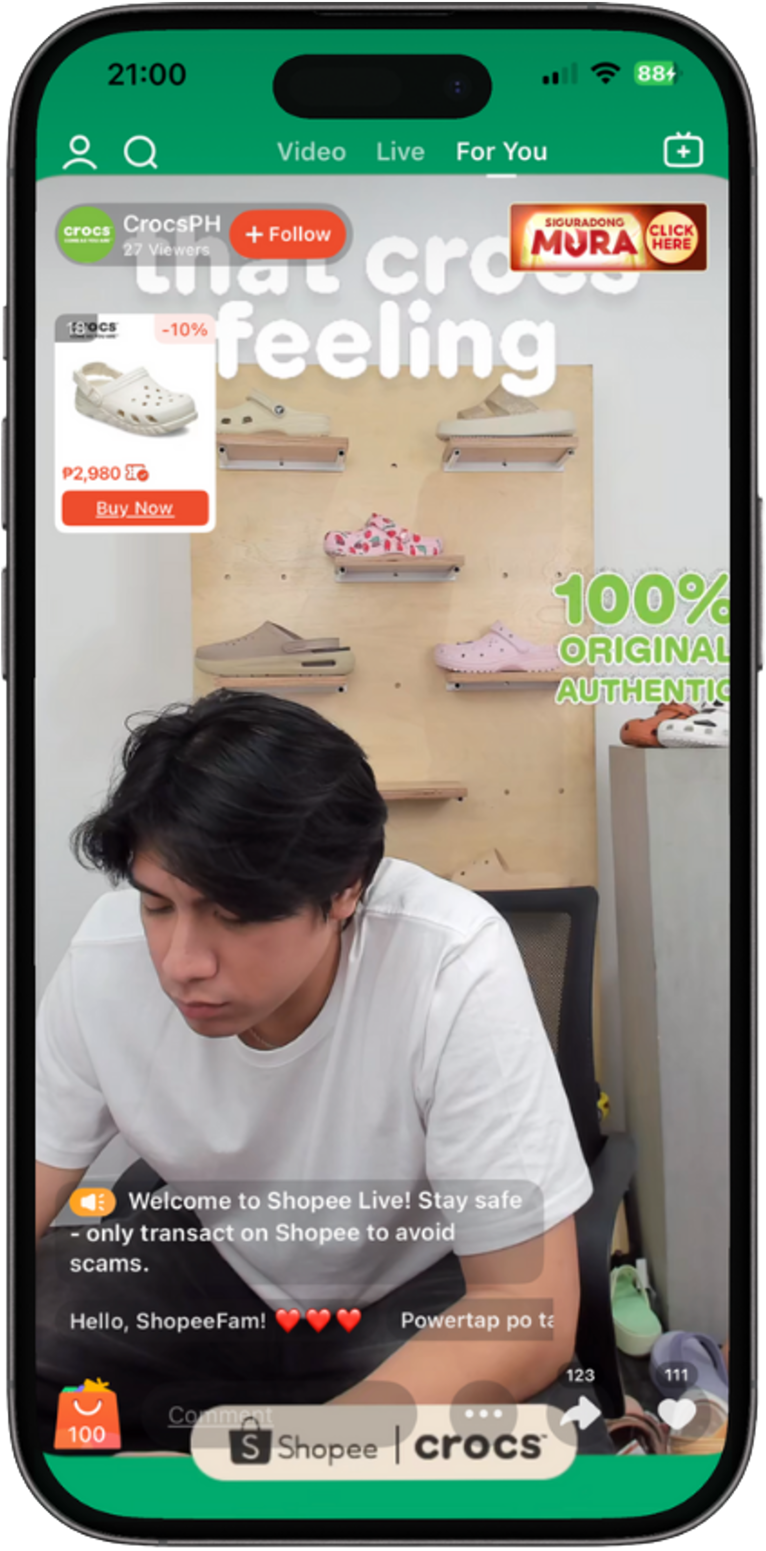 A person shown on a mobile device screen during a Shopee Live shopping stream, featuring Crocs shoes display in the background and an advertisement for Mura. The user interface includes icons for live video, for you, and follow options, and a shopping section with a Crocs shoe promoted at 10% off.