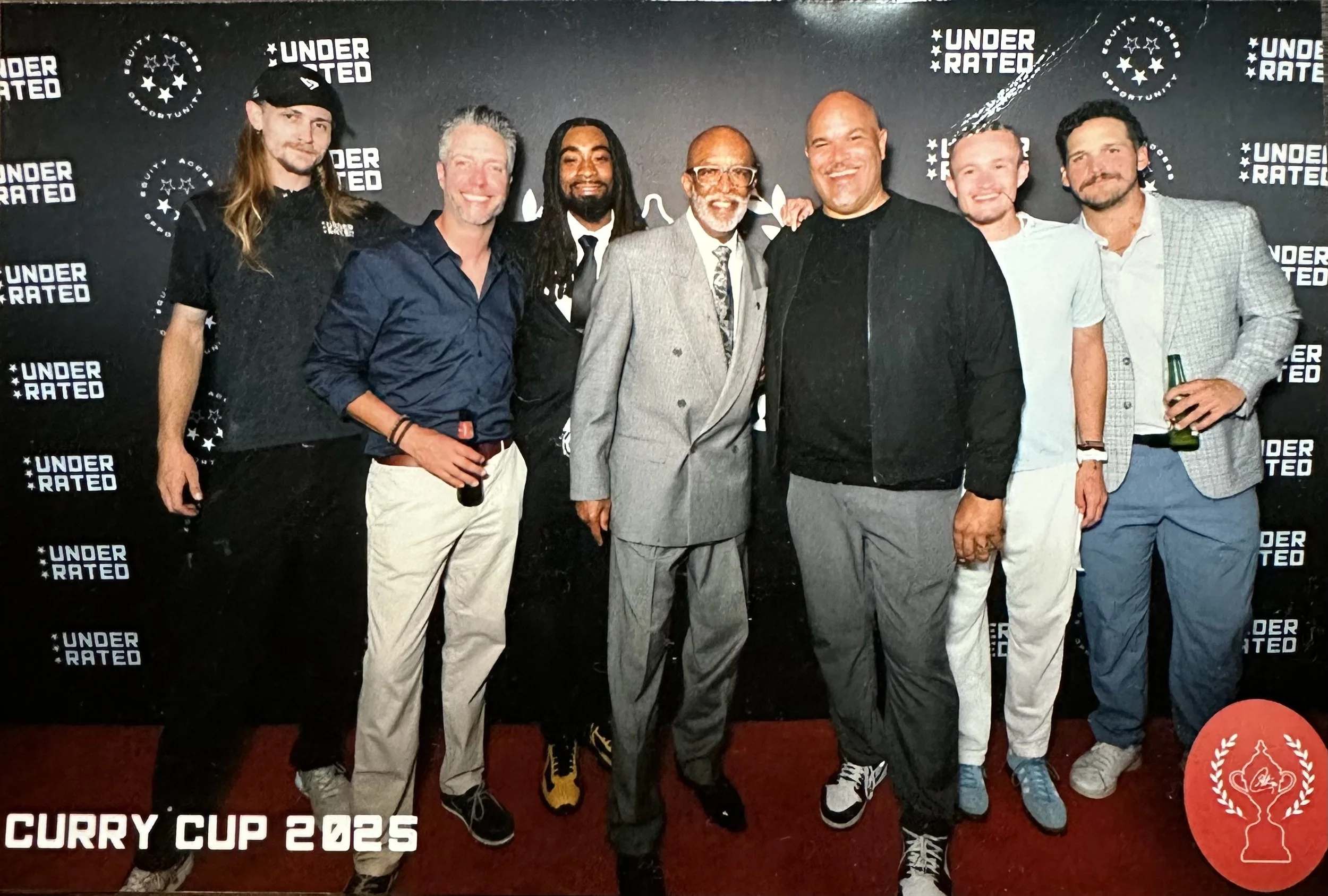 Group of eight men posing together on a red carpet at the Curry Cup 2025 event, with a backdrop displaying the event's logo and the words 'Under Rated.'