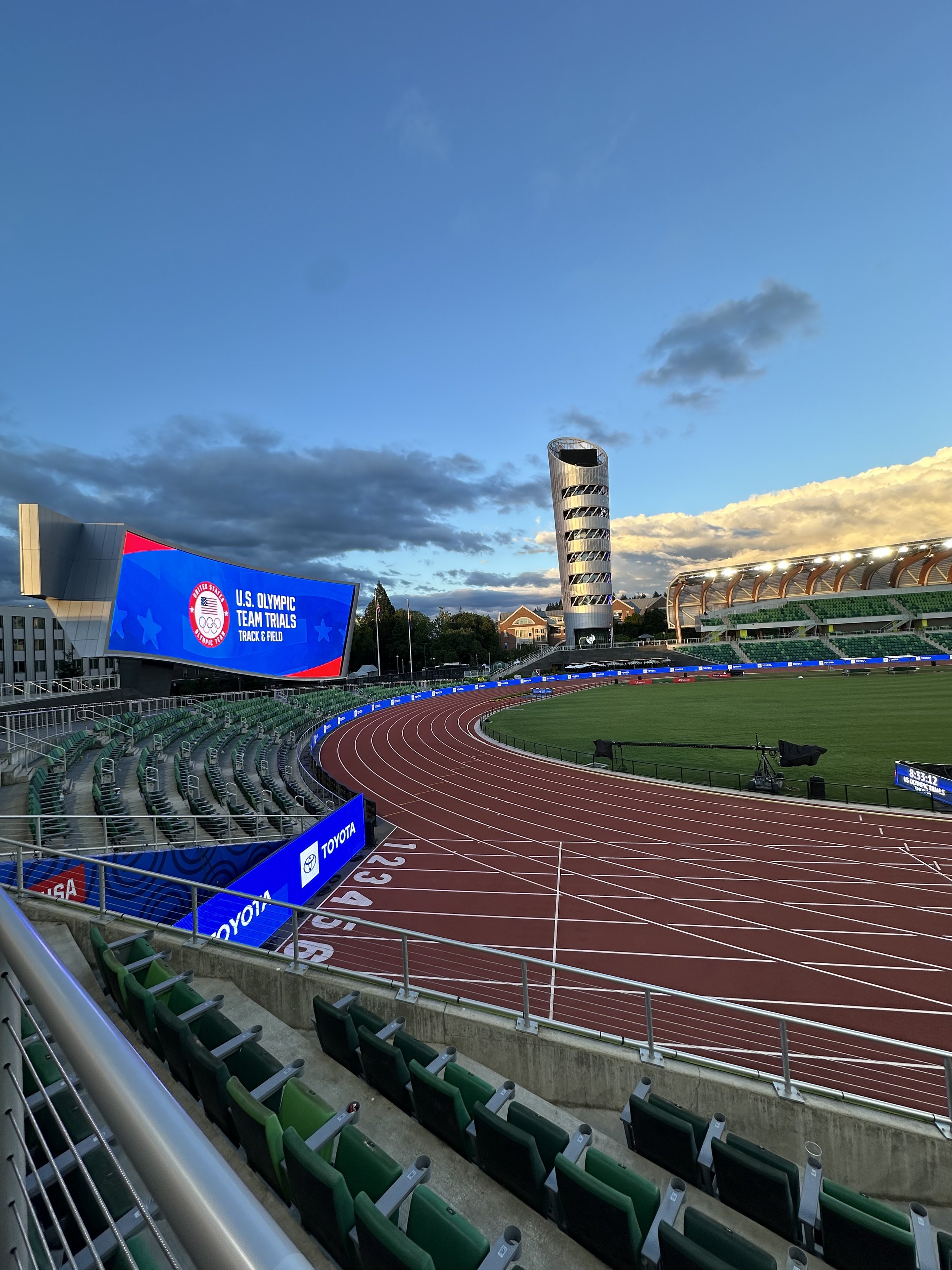 An empty outdoor stadium with a running track, green seats, and digital screens displaying the U.S. Olympic Team Trials for track and field, under a sky with clouds.
