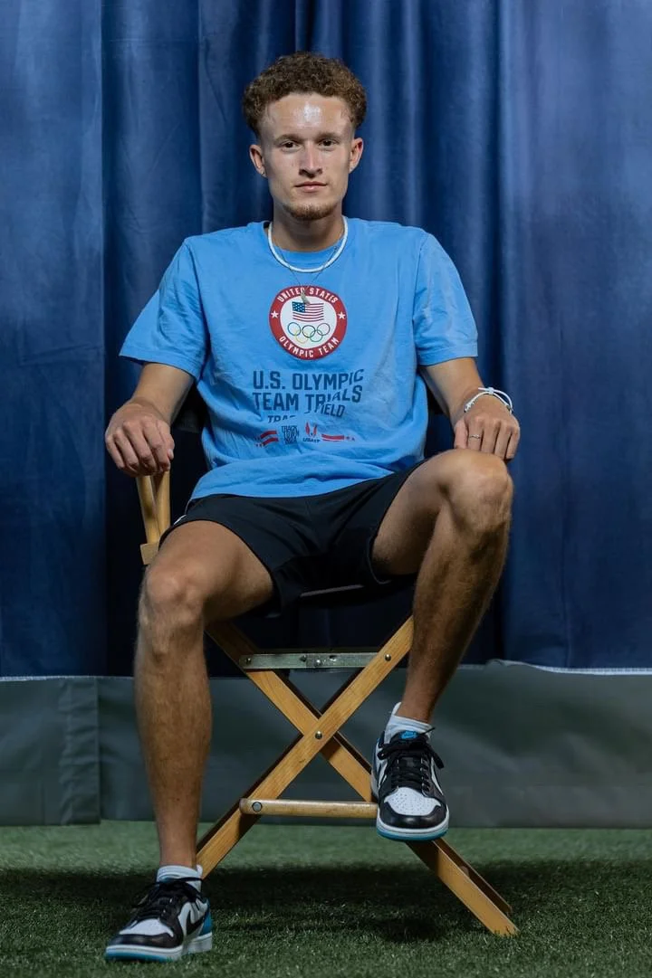 Young man sitting on a wooden chair in front of blue curtain, wearing a blue U.S. Olympic Team Trials T-shirt, black shorts, and athletic shoes.