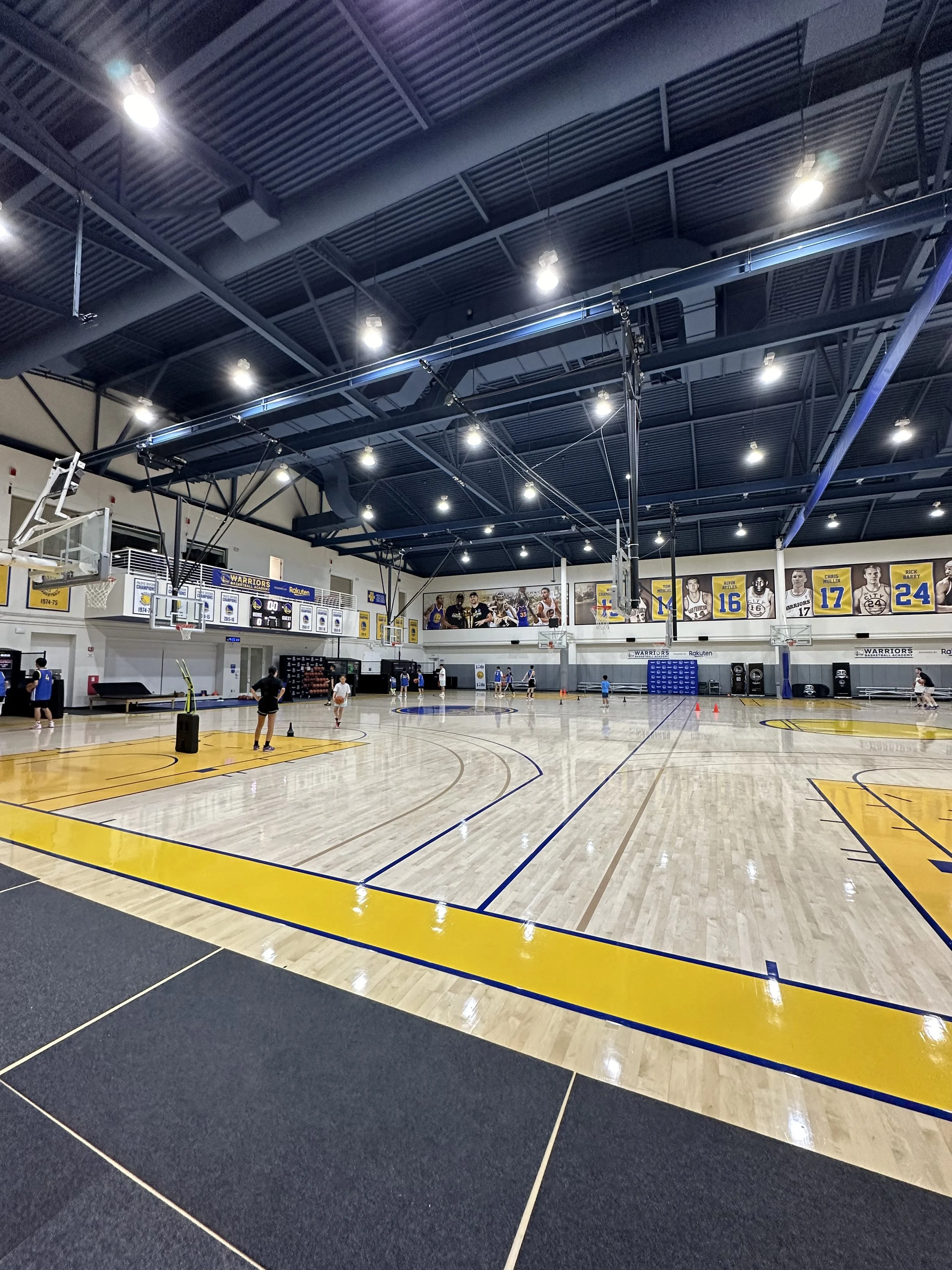 Indoor basketball court with players practicing, surrounded by banners and photos of basketball players on the wall, brightly lit with ceiling lights.