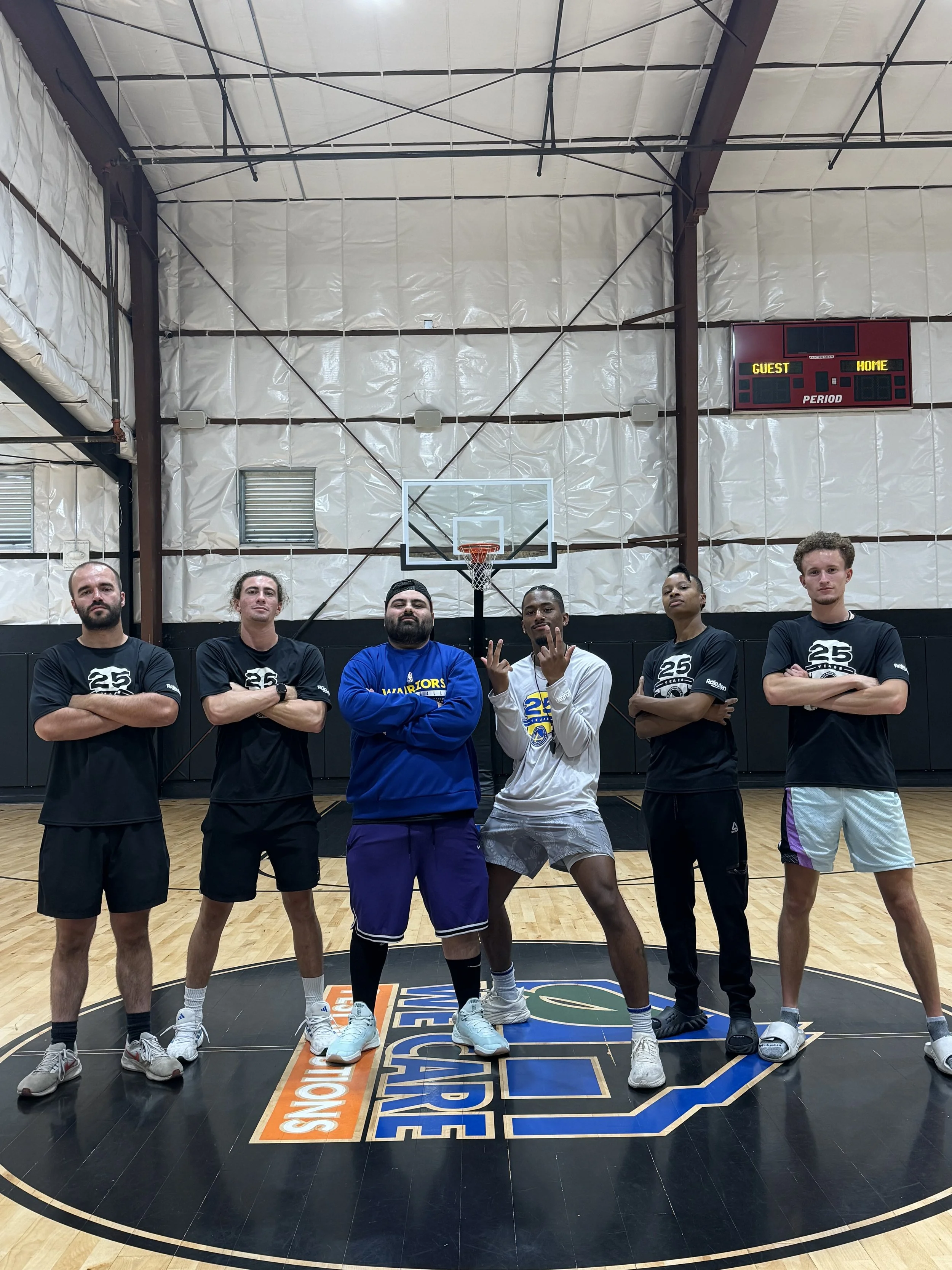 Group of six basketball players standing inside an indoor court with arms crossed, posing on the court's logo, some wearing Warriors apparel.