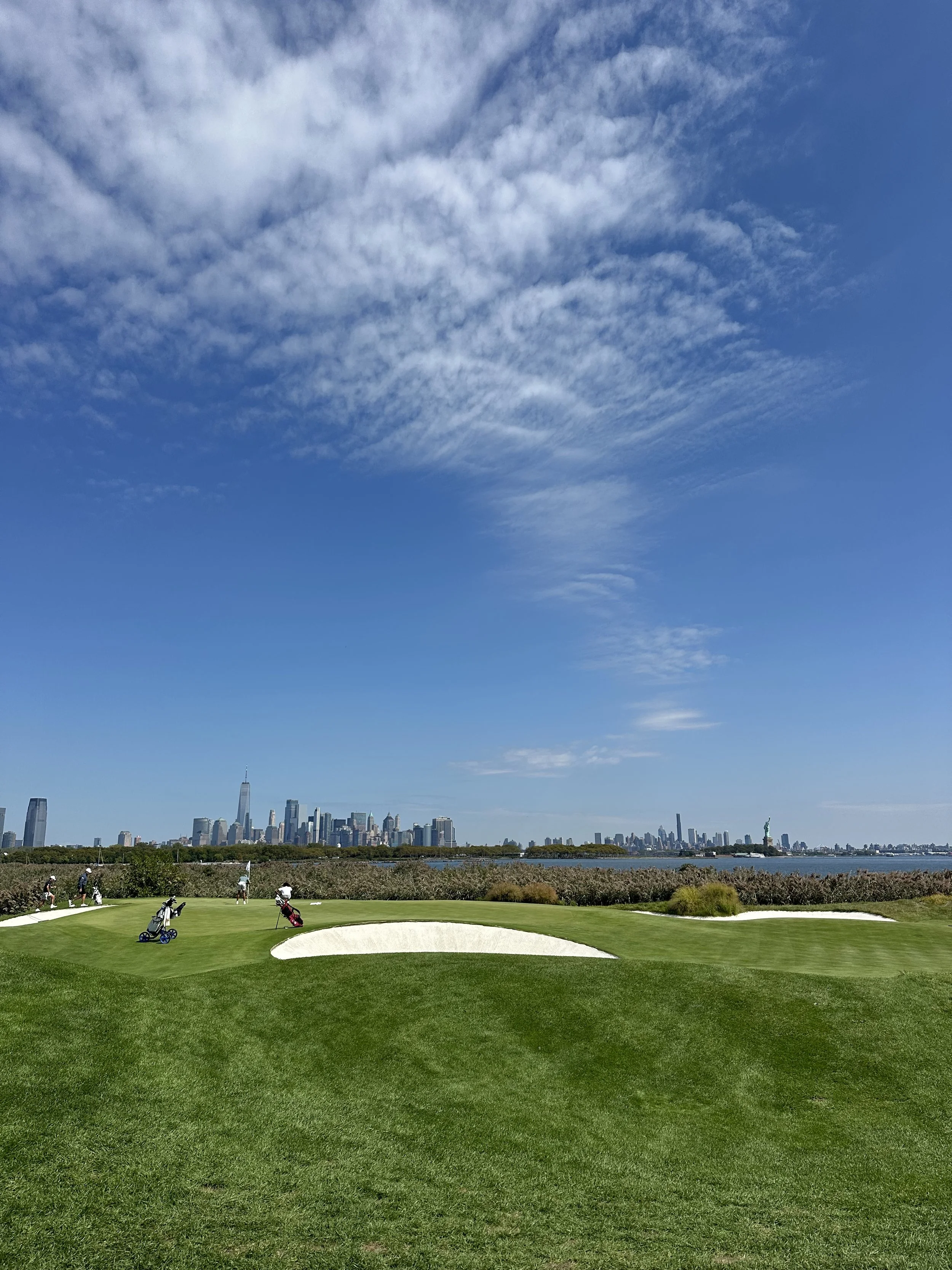Golf course with sand traps and players, New York City skyline in the background, clear blue sky with scattered clouds.