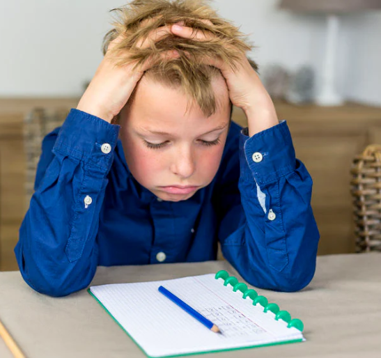 Child sitting at a table with a notebook and a pen, looking frustrated with hands on his head.