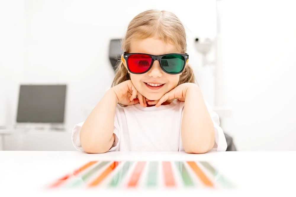 A young girl wearing 3D glasses with red and green lenses smiling and resting her chin on her hands, sitting at a table with an anaglyph in front of her.