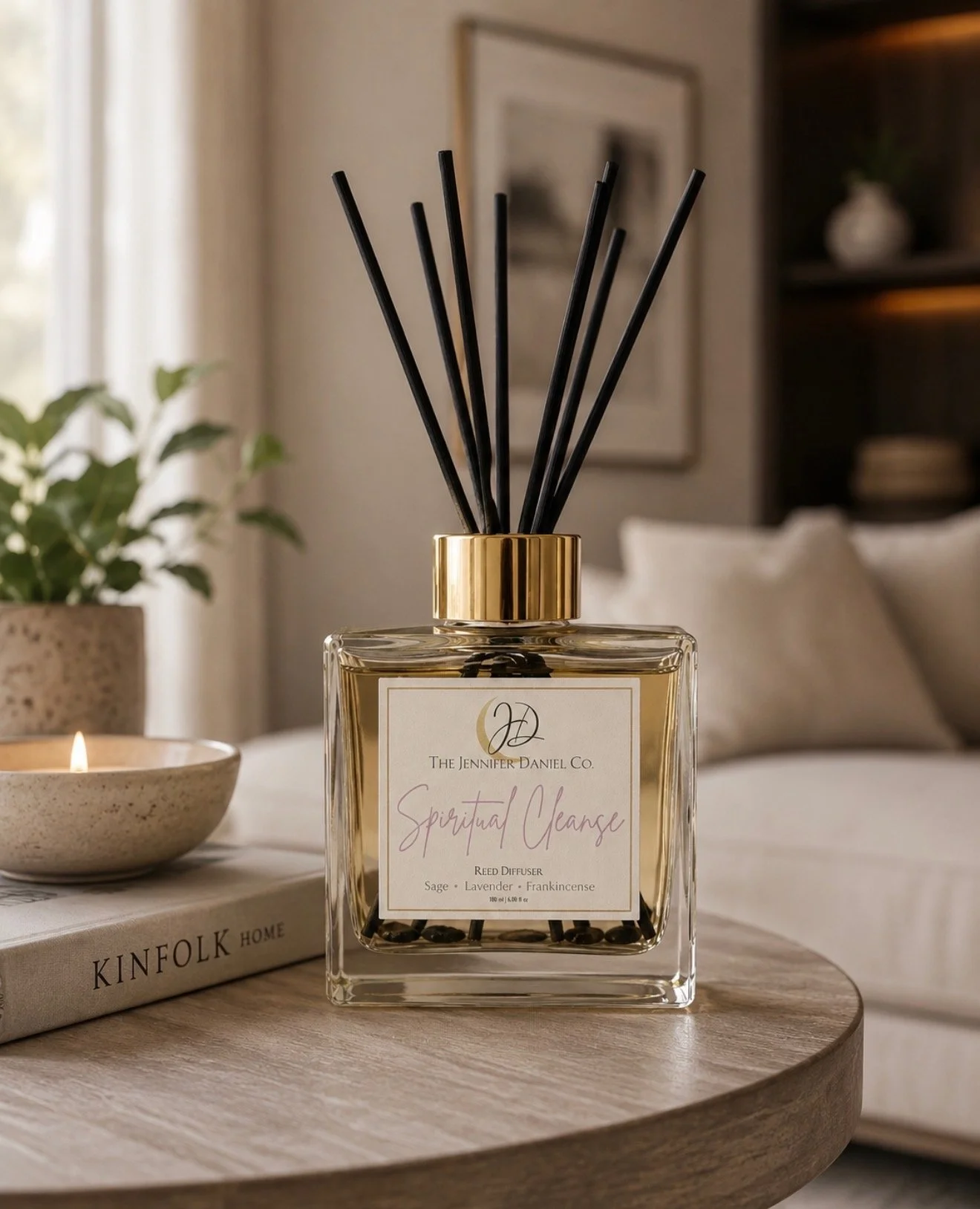 A reed diffuser with black reeds and a gold cap, labeled "Spiritual Cleansing" with scents like sage, lavender, and frankincense, placed on a wooden table alongside a lit candle and a book titled "Kinfolk Home" in a cozy, softly lit room.