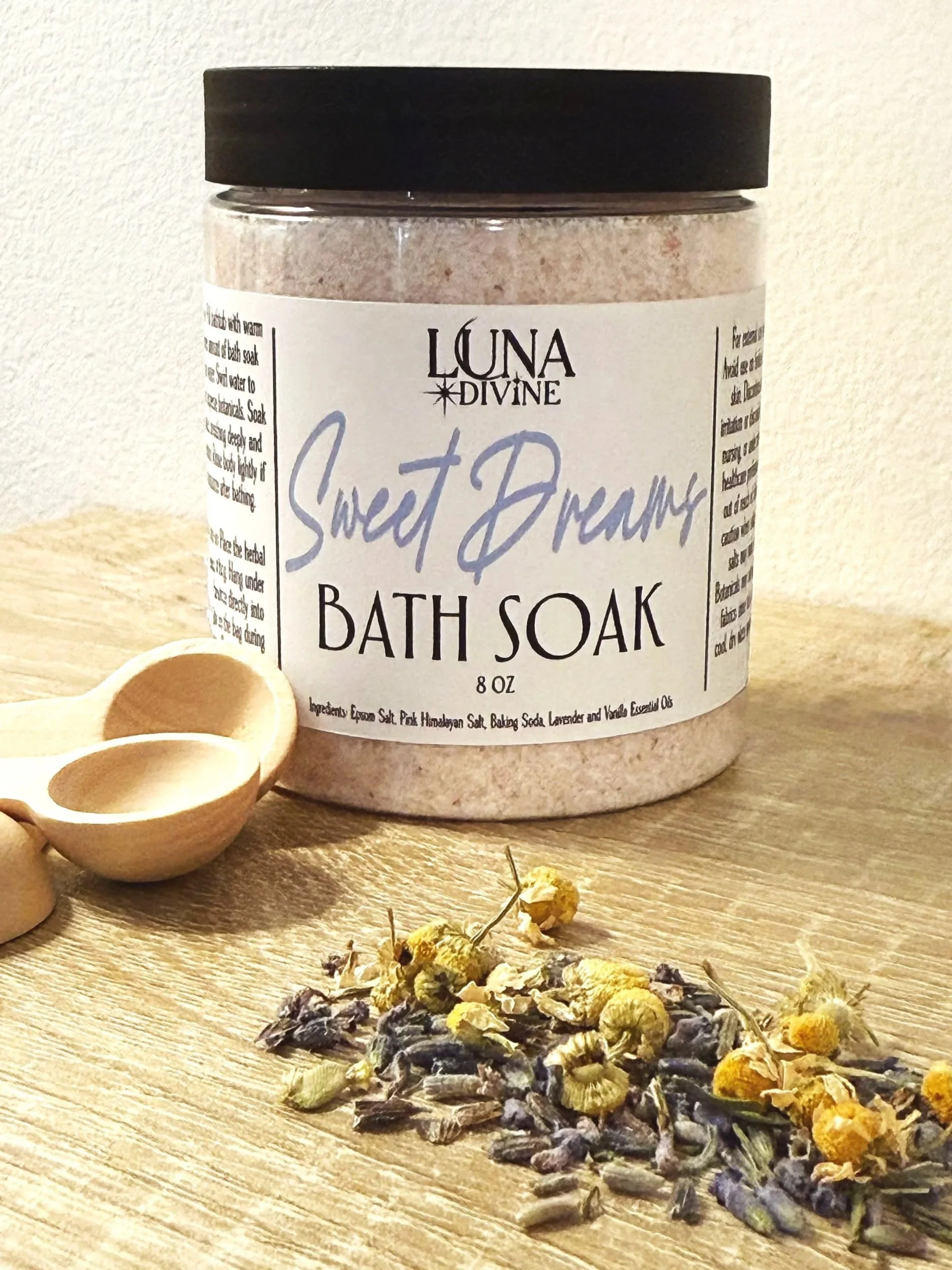 A jar of Luna Divine Sweet Dreams bath soak with dried lavender and chamomile flowers in front, along with two small wooden scoops on a wooden surface.