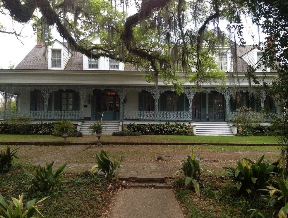 A large, historic house with Victorian architecture, featuring a wide porch, intricate ironwork, and white stairs leading up to it. The house is surrounded by green lawn and trees with hanging moss.