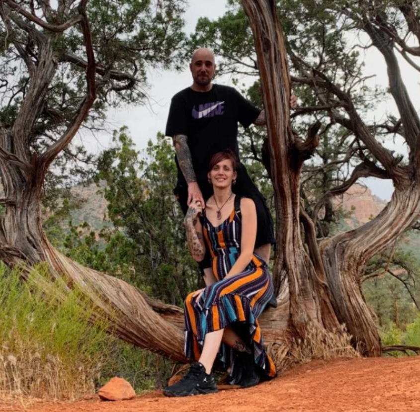 A woman sitting on a tree branch with a man standing behind her, holding the branch, in a natural outdoor setting with trees and red dirt.