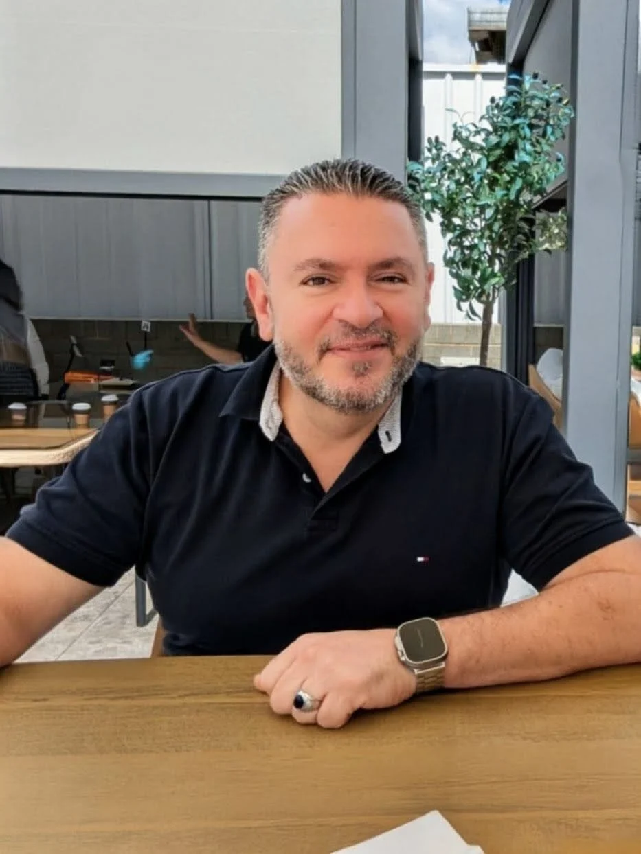 A middle-aged man with short, slicked-back hair and a beard, wearing a black polo shirt and a smartwatch on his left wrist, sitting at a wooden table in a modern outdoor dining area with a small potted tree and gray walls.