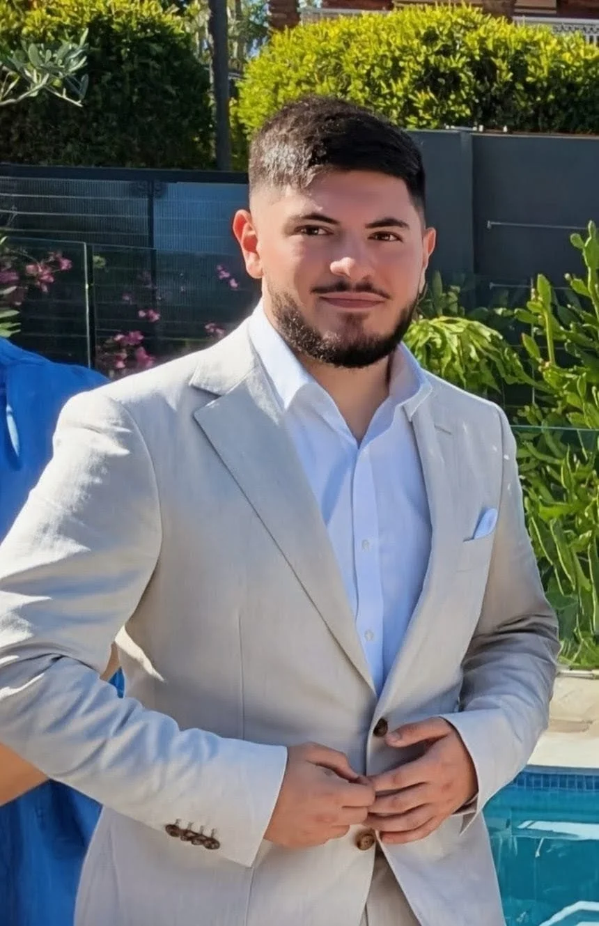 A young man with dark hair and a beard is wearing a light-colored suit jacket over a white dress shirt, standing outdoors near a pool and greenery.