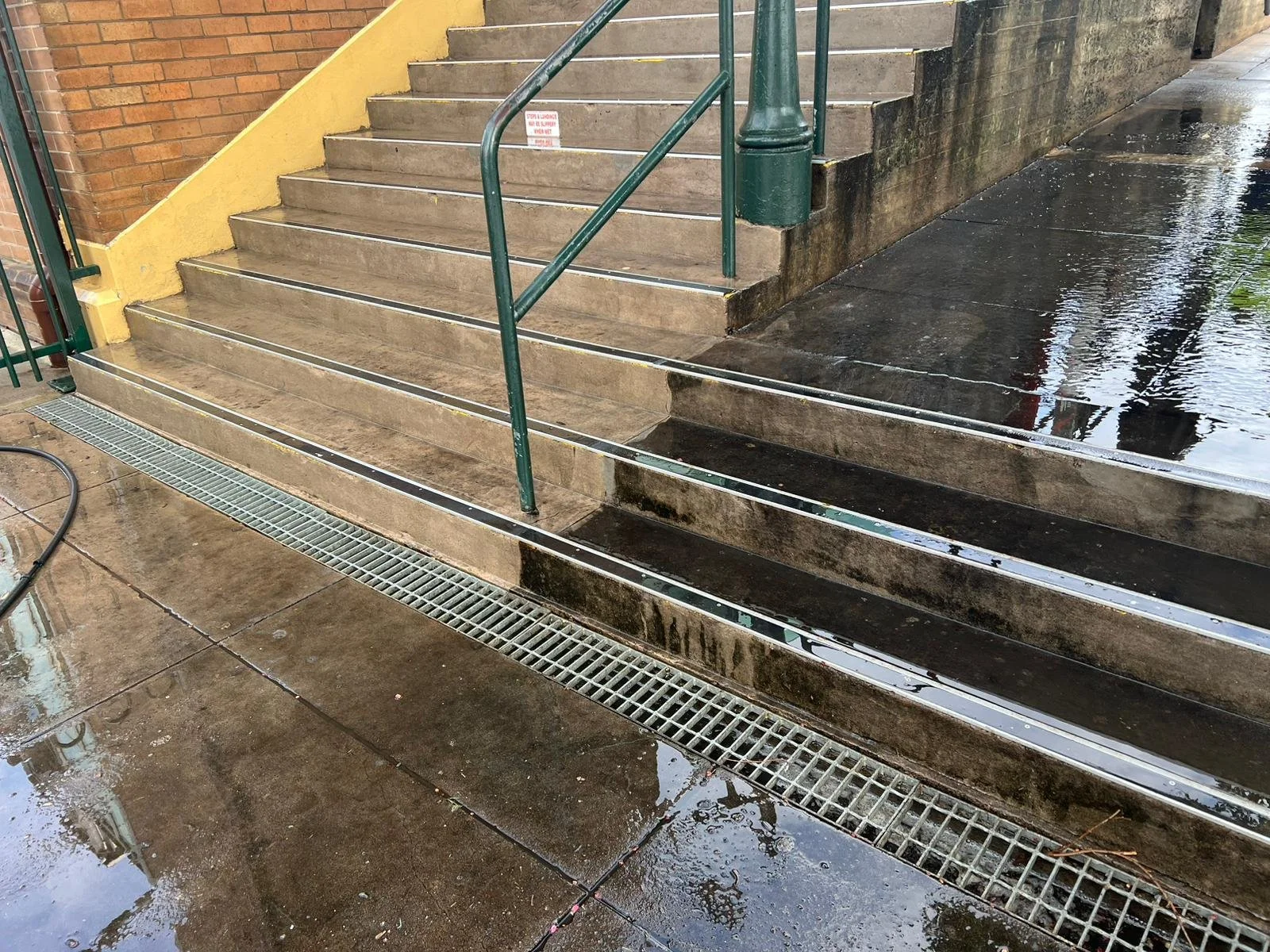 Rainy day at a staircase with a ramp for accessibility, with wet concrete and drains on the ground, and a yellow wall on the side.