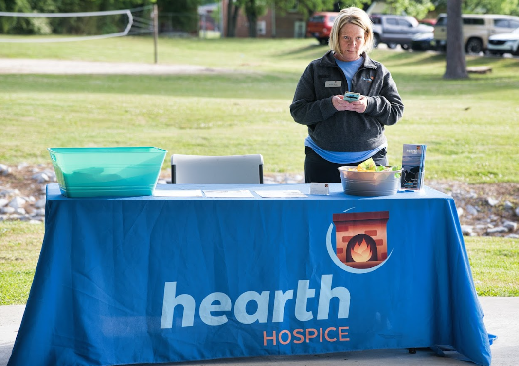 A woman standing behind a blue tablecloth with the Hearth Hospice logo, outdoors on a grassy area, with a bowl of snacks and informational flyers on the table.