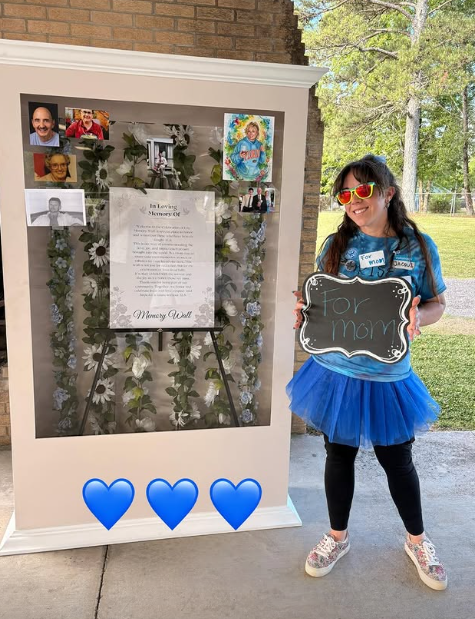 A girl wearing sunglasses and a blue tutu stands next to a memorial display with photographs and a message, holding a small chalkboard sign that says 'For mom.'