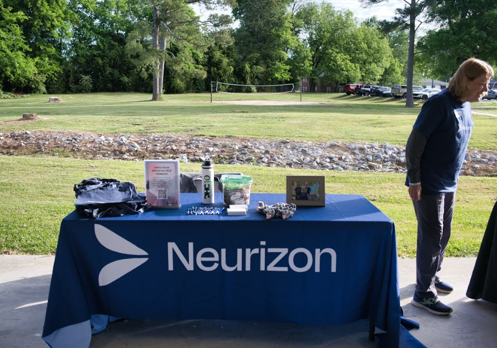 Outdoor event setup with a blue Neurizon tablecloth, informational materials, and a person standing beside the table on a grassy area with trees and parked cars in the background.