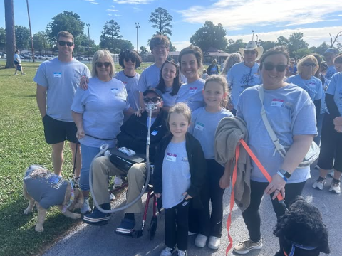 Group of people, including children and adults, standing together outdoors on a sunny day. Some are wearing light blue T-shirts, and there are dogs with leashes. The background shows trees, a parking lot, and other groups of people.