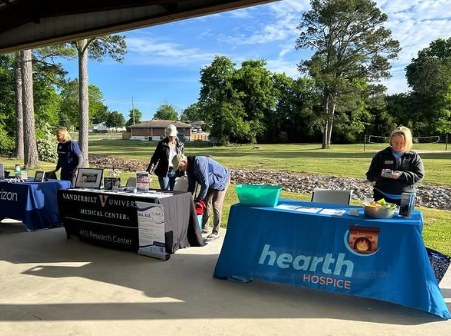 People at outdoor informational booths at a community event under a pavilion, with trees and grassy area in the background.