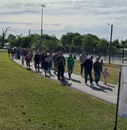 A long line of diverse people, including children, walking along a sidewalk next to a grassy area and a fenced sports court on a partly cloudy day.