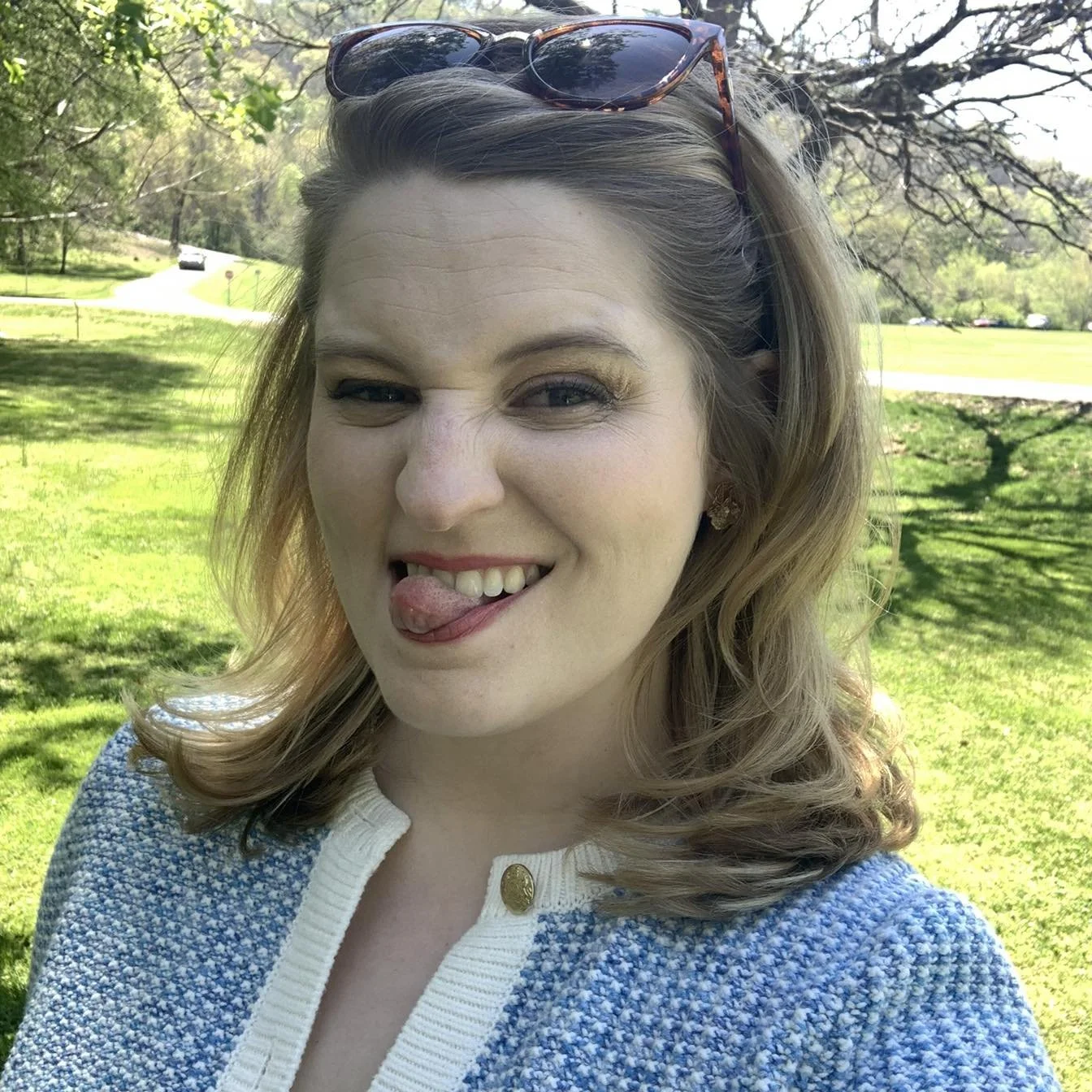 A young woman with wavy, shoulder-length hair, sunglasses resting on her head, sticking out her tongue and winking in a park on a sunny day.