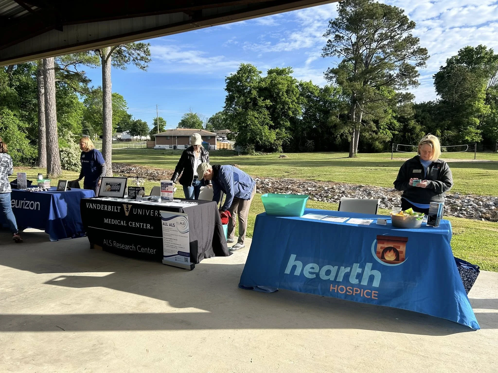 Outdoor event with tables and banners from Vanderbilt University Medical Center, Heart Hospice, and Verizon, under a pavilion with trees and a volleyball net in the background.
