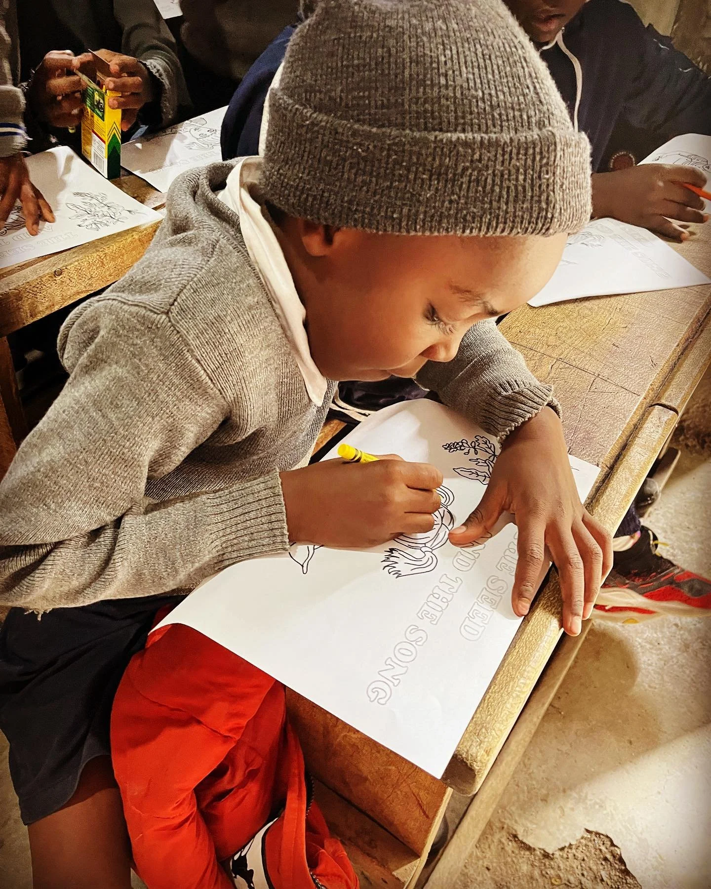 A young boy wearing a gray beanie and a gray sweater is sitting at a wooden desk, coloring a large printed poster with the words 'SCORED REBEL SO' and a drawing of a lion.