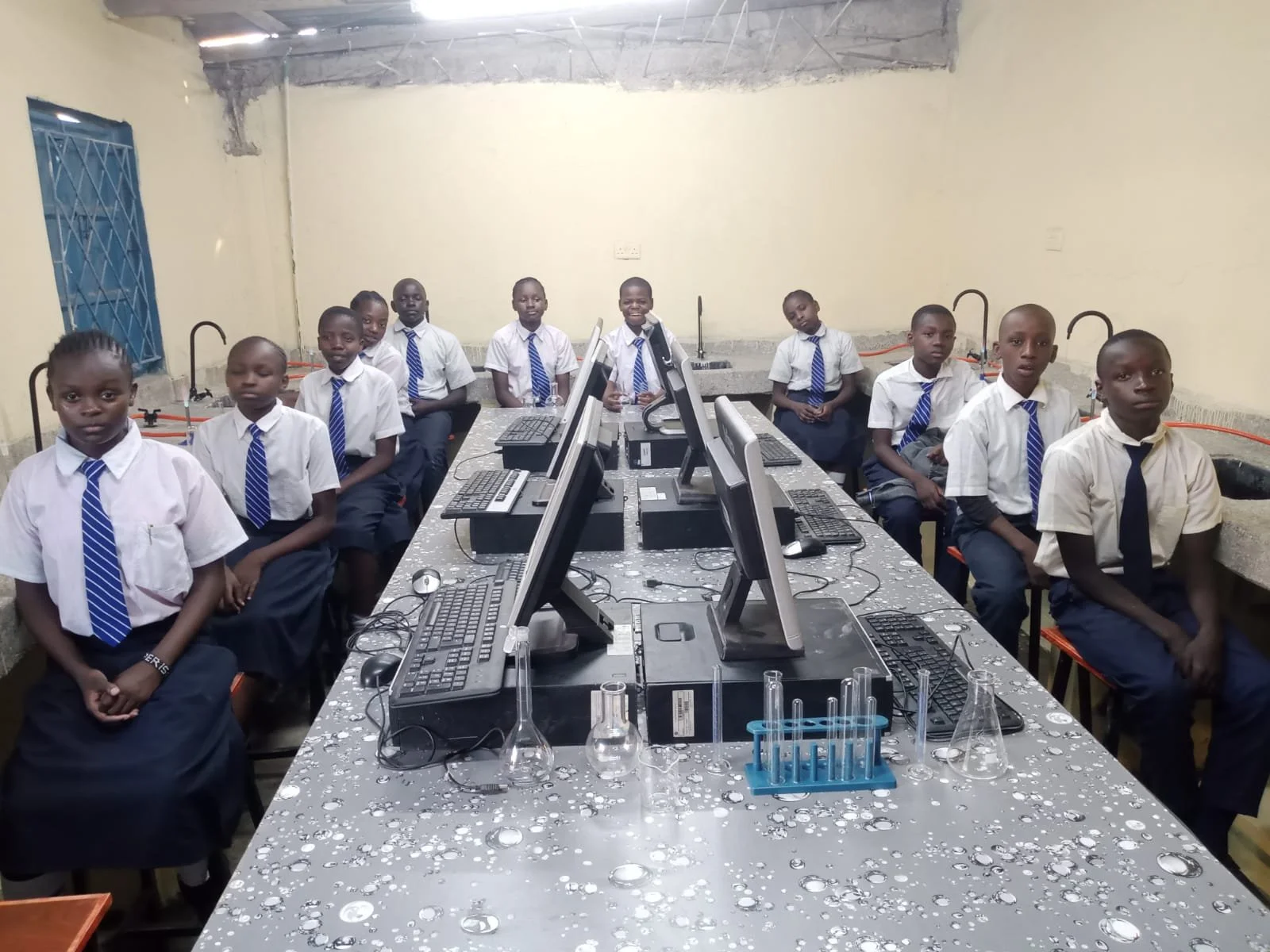 A classroom with students wearing school uniforms seated around a table with computers and laboratory glassware.