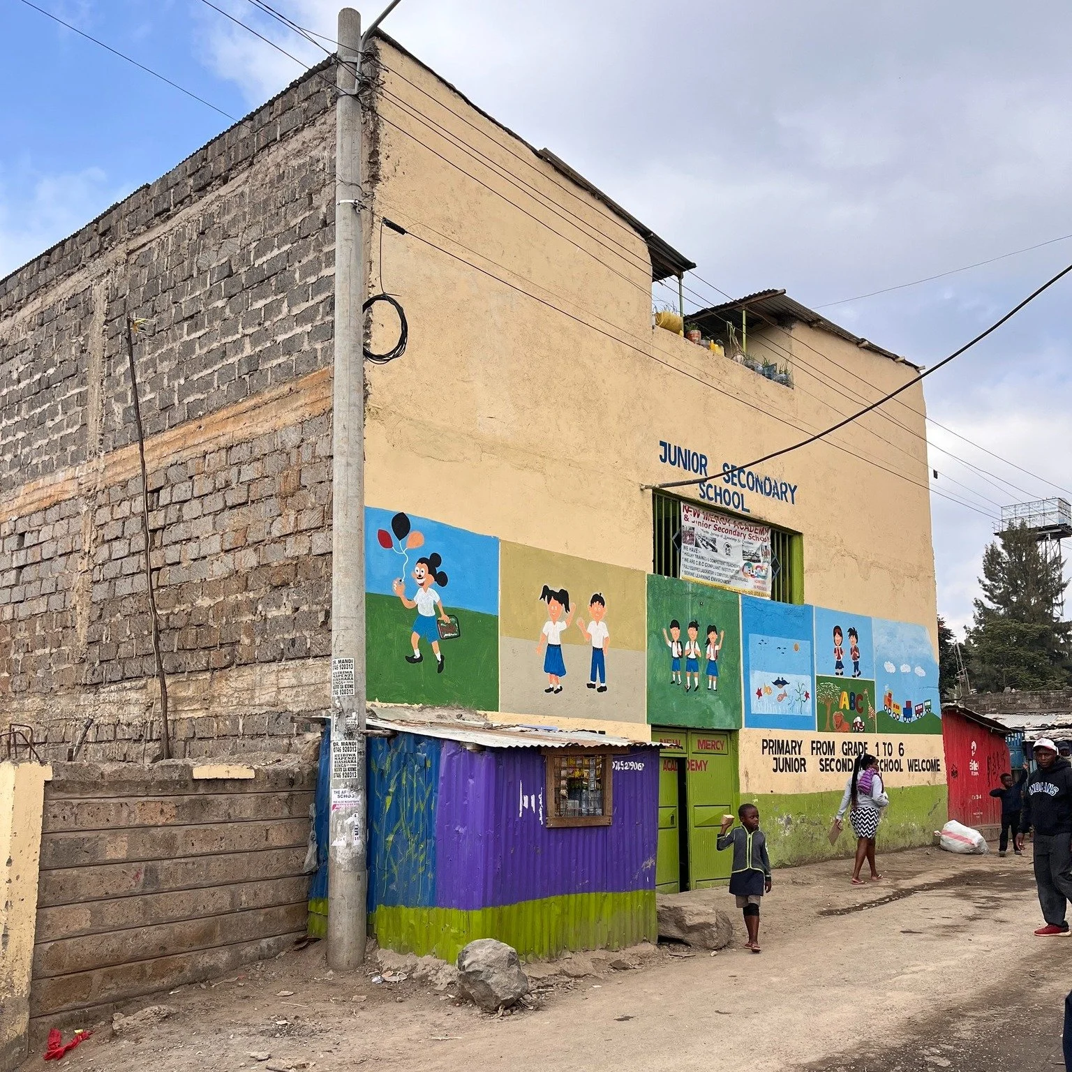 A two-story building with a painted mural of children on the wall, labeled 'Junior Secondary School,' with people, including children and adults, walking nearby on a dirt path.