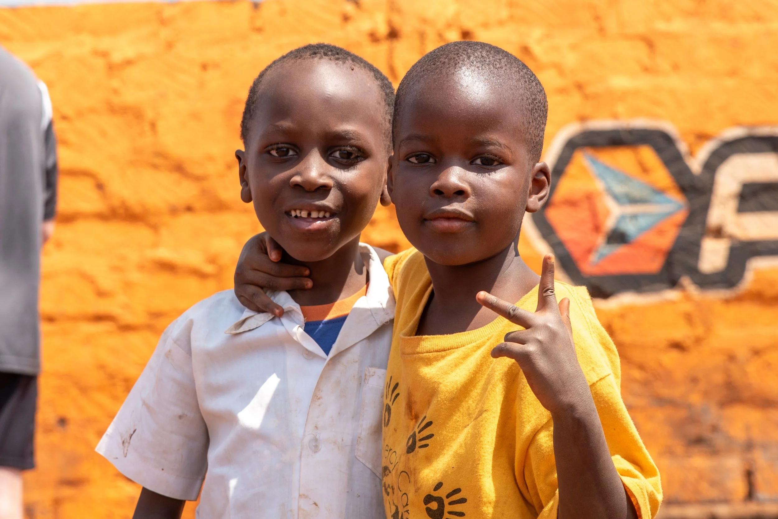 Two young boys standing close together, one with arm around the other's shoulder, posing for a photo outdoors against a bright orange wall with a colorful geometric mural.