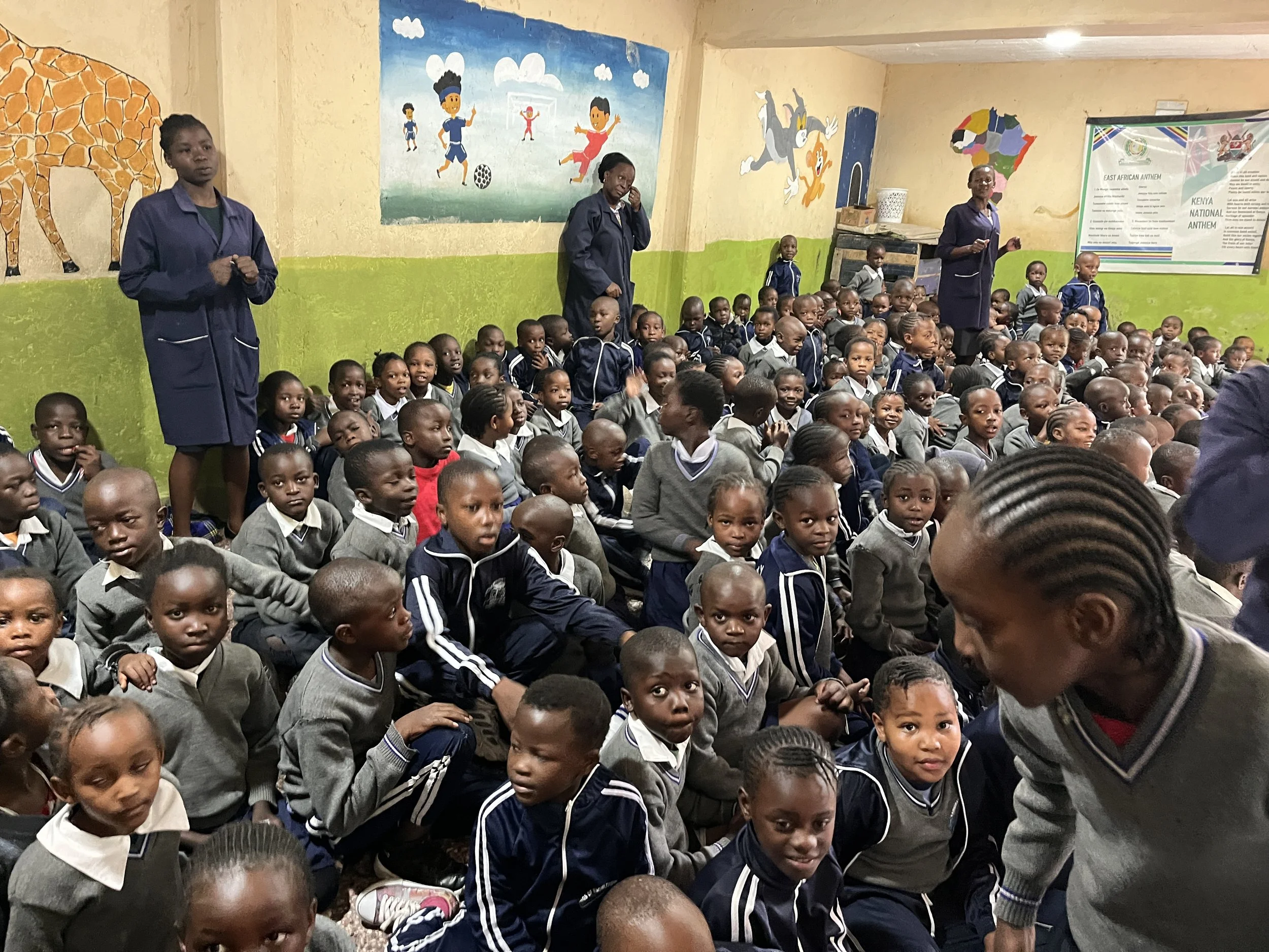A large group of young African school children seated on the floor in a classroom, with some standing, supervised by teachers or staff, with colorful wall paintings and a Kenyan map and anthem poster in the background.