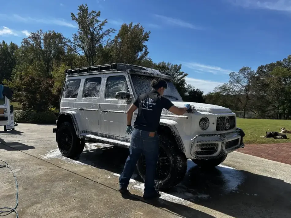 A man cleaning a white SUV with soap and water outside on a sunny day.
