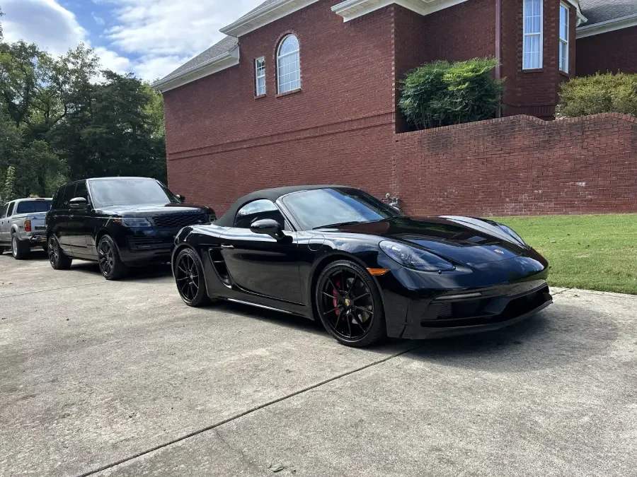 A black sports car, possibly a Porsche, parked on a driveway in front of a brick house, with a black SUV and a silver truck parked behind it.