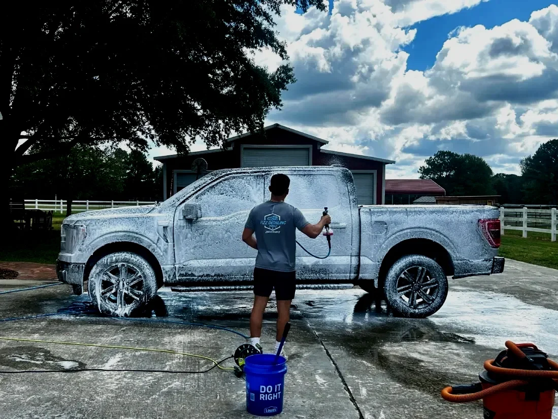 A person washing a large pickup truck with soap and a hose outside on a concrete driveway, with a barn in the background and clouds in the sky.