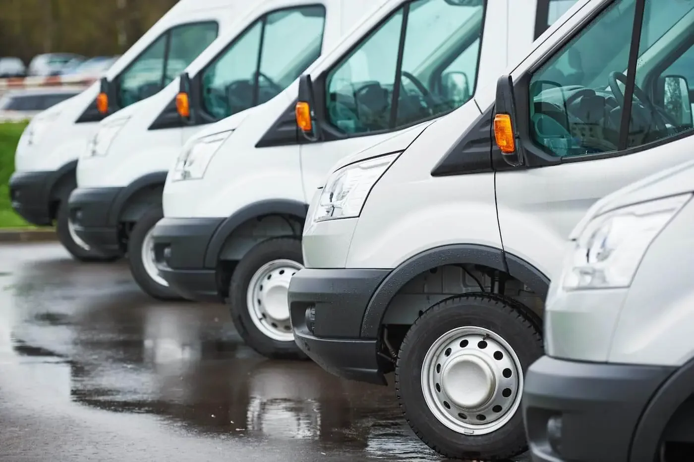 Multiple white commercial vans parked in a row on a wet surface.