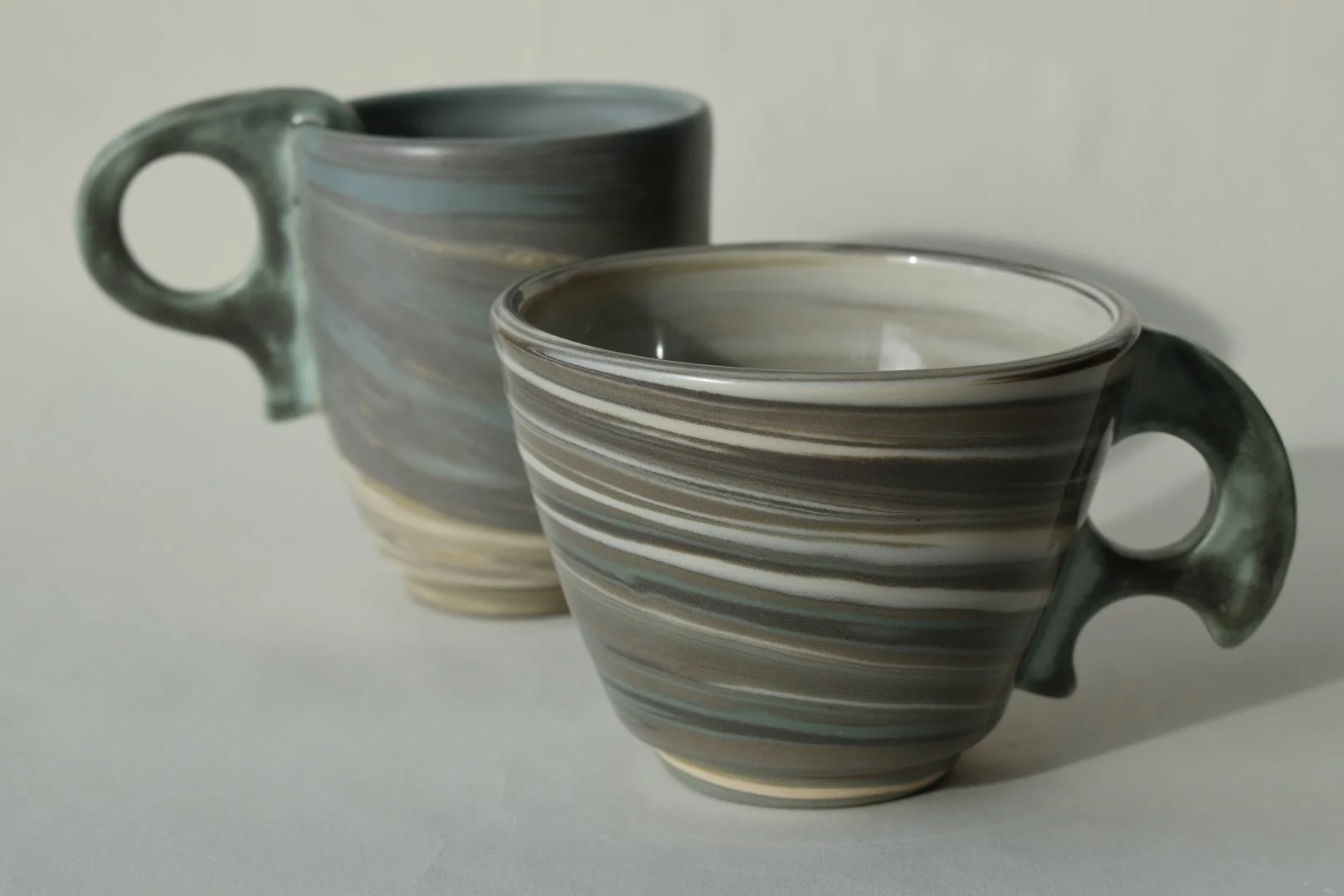Two ceramic cups with striped patterns, one in the foreground and one in the background, both with loop handles, placed on a flat surface.