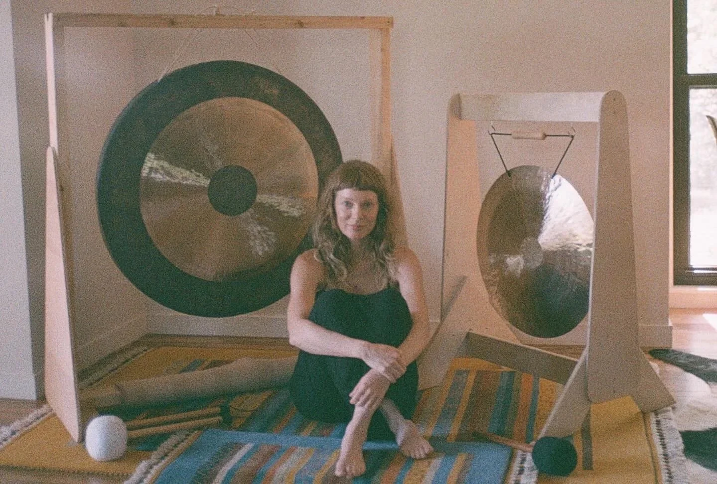 A woman sitting on a rug indoors, surrounded by large gongs, with one gong mounted in a wooden frame and another hanging from a stand.