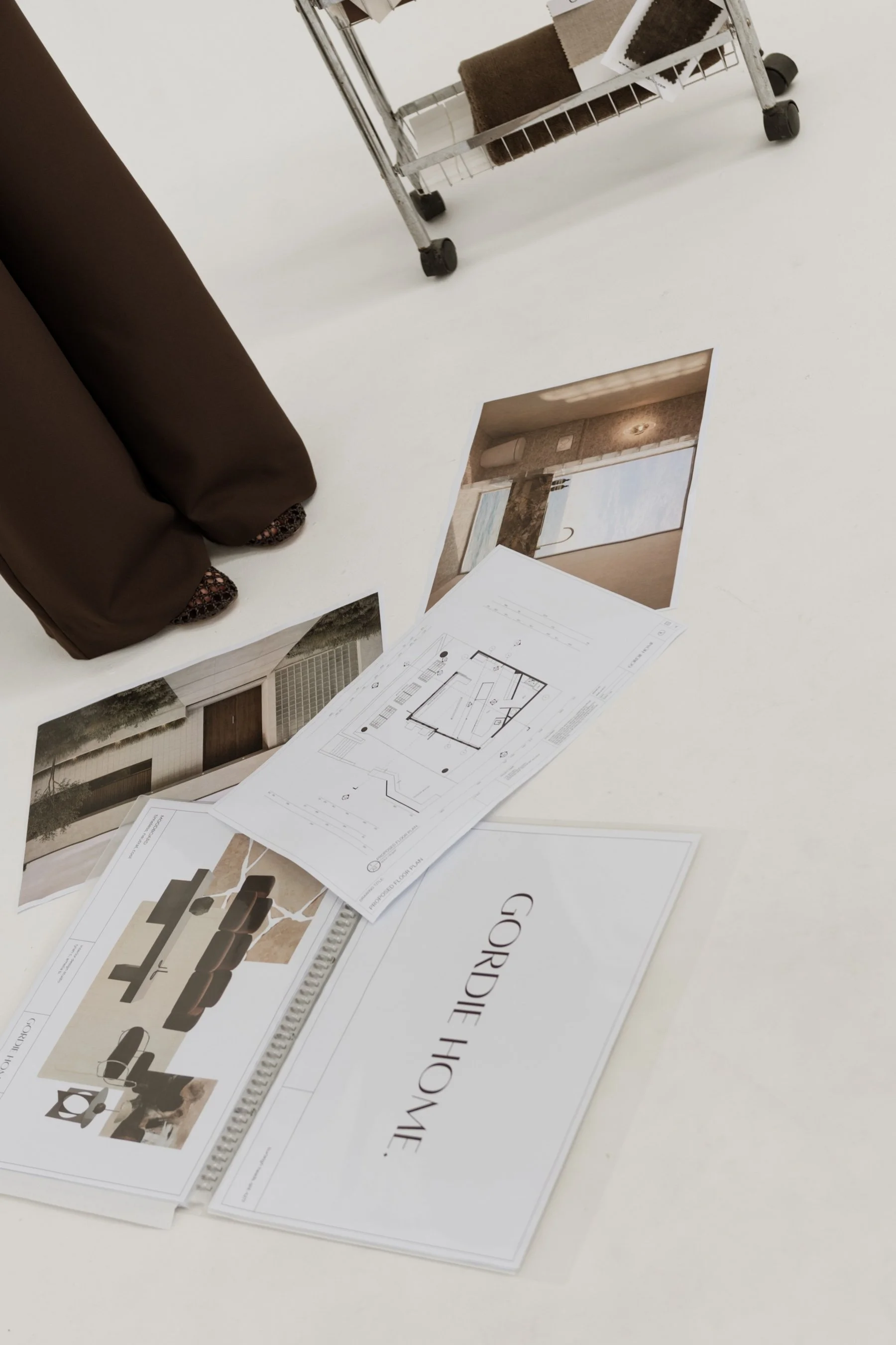 Design plans, photos, and material samples labeled 'GORDIE HOME' arranged on a white table, with a metal cart holding additional samples in the background.